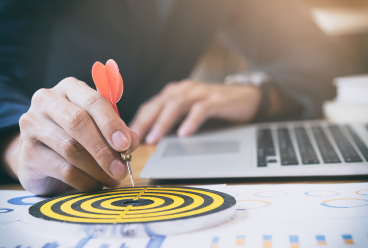 A person is holding a dart ready to aim at the bullseye of a target, with a laptop and business charts in the background, symbolizing goal setting and achievement in a business context.